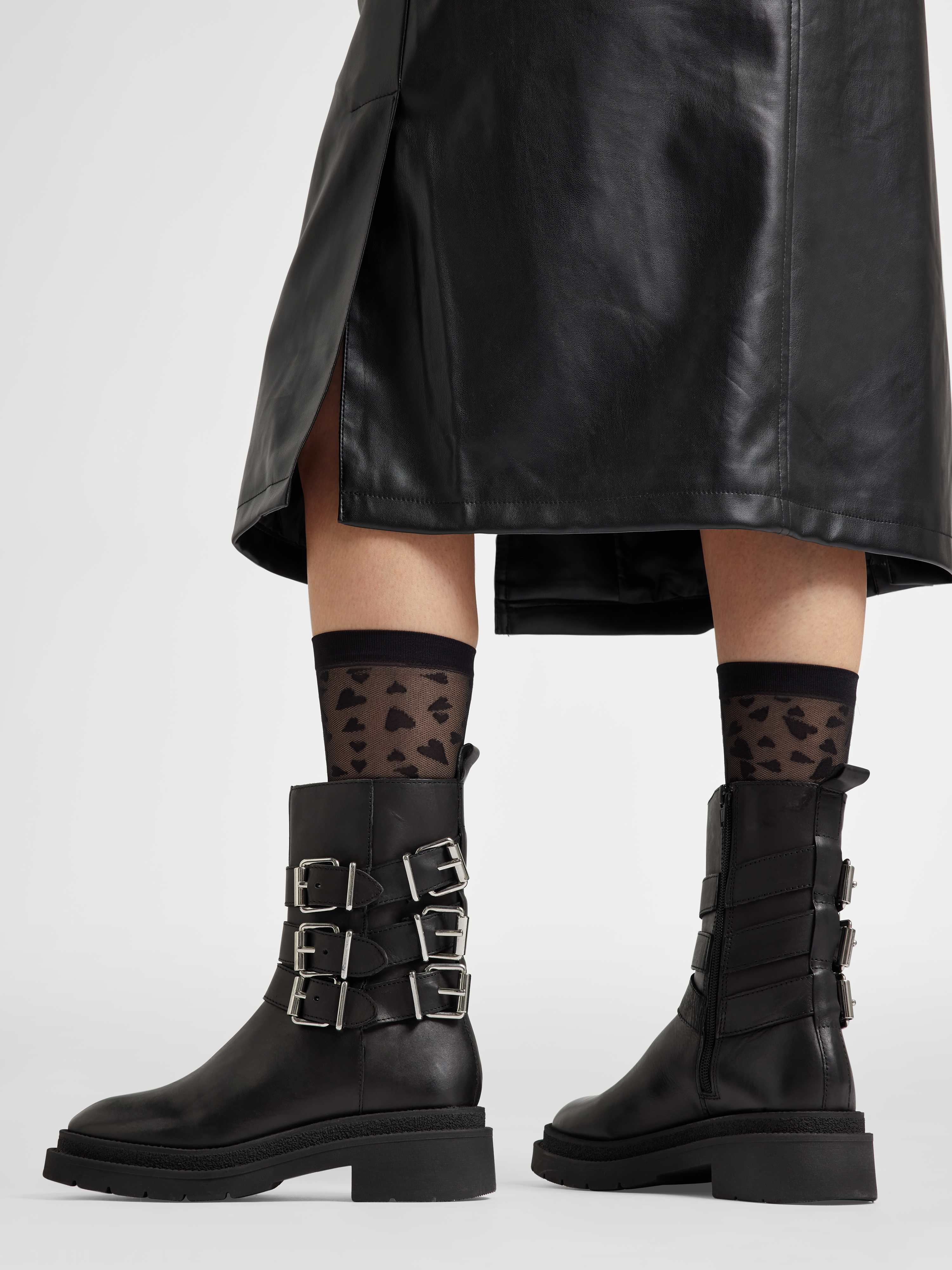 Person trägt schwarze Biker-Boots mit Metallschnallen, vor weißem Hintergrund