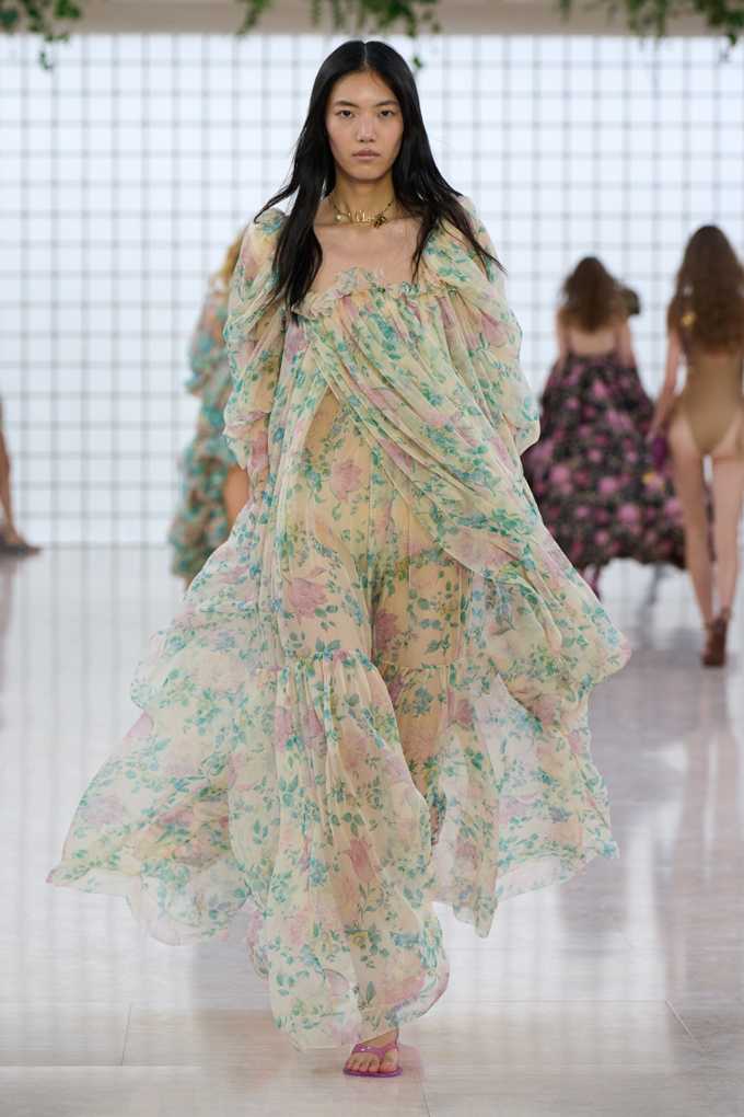 Frau mit langen Haaren und weitem Kleid mit Blumenmuster, gehend auf einem Laufsteg