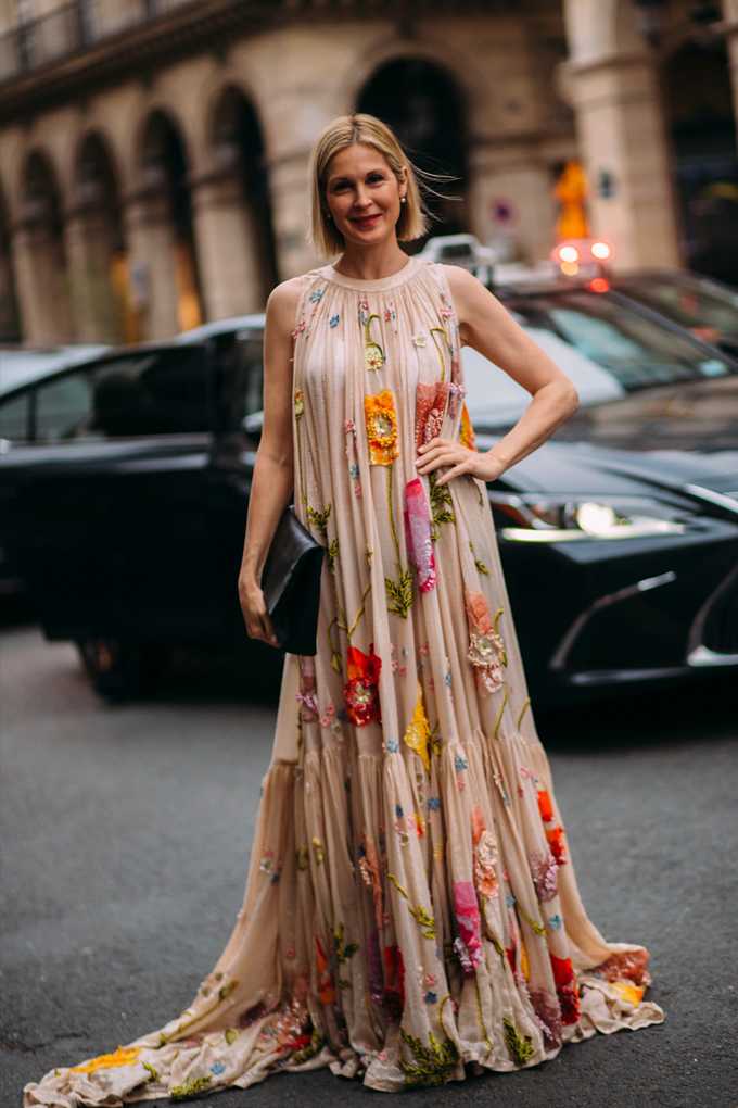 Frau in leichtem Kleid mit auffälligen Blumen-Stickereien, Hand in Hüfte gestützt, stehend auf Straße
