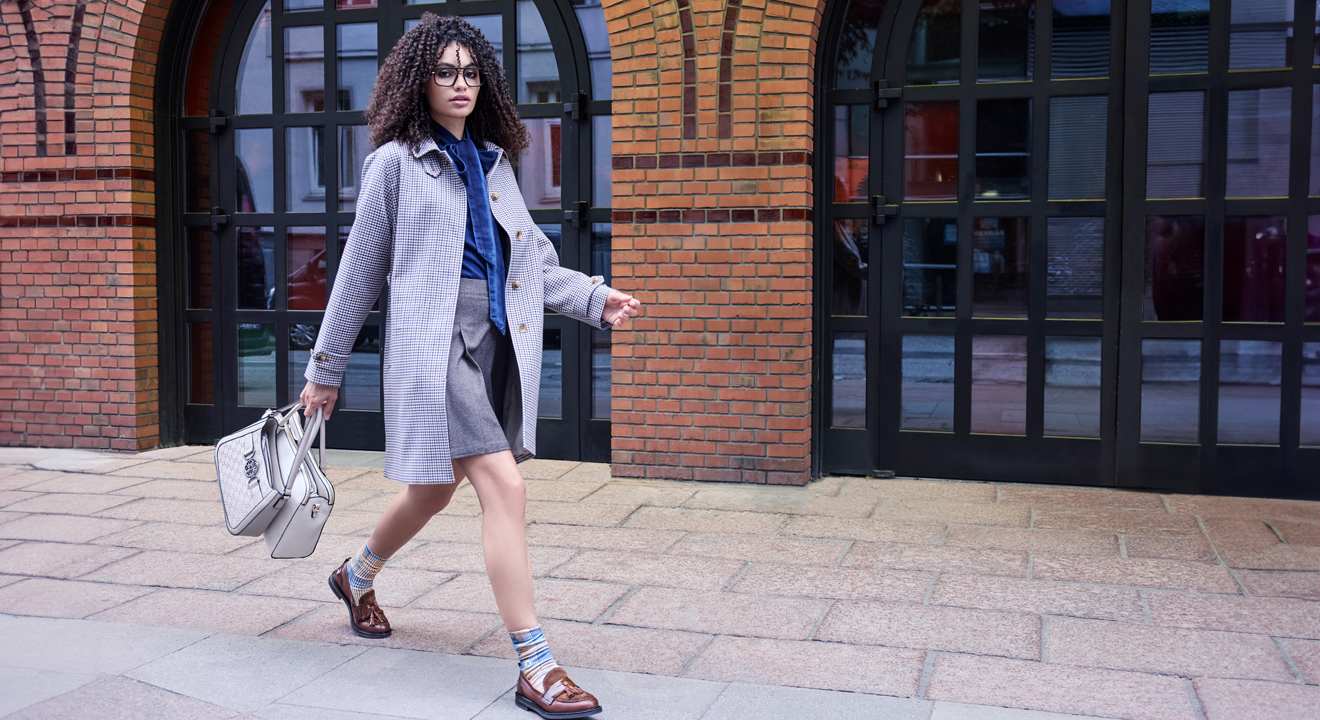 Frau in kariertem Mantel, blauem Hemd, grauem Rock und braunen Loafern mit einer weißen Tasche