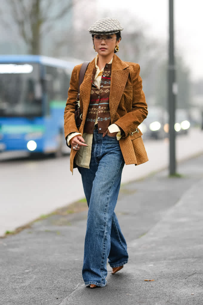 Frau spaziert in warm-braunem Blazer, trägt blaue Jeans und Schiebermütze