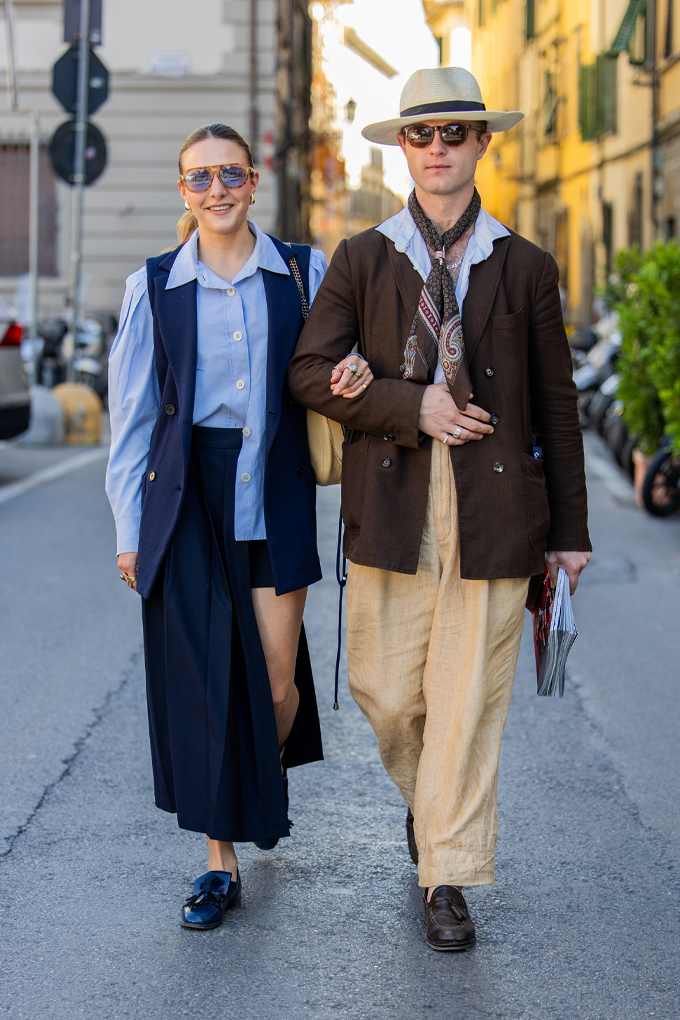 Ein Paar läuft auf einer Straße. Die Frau trägt eine Bluse, Weste und Rock. Der Mann trägt Hut, Sonnenbrille, Hemd, Krawatte, Jacke und Hose.