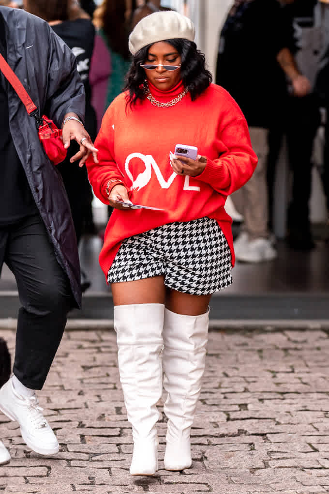 Eine Frau trägt beiges Barett, Sonnenbrille, einen in die Hose gesteckten roten "LOVE"-Pullover, schwarz-weiß karierte Shorts und weiße Stiefel, während sie geht und auf ihr Handy schaut.