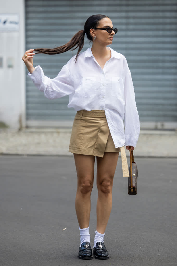 Frau trägt weißes Oversize-Hemd teils in beigefarbene Minirock gesteckt kombiniert mit Loafern, Socken und Sonnenbrille