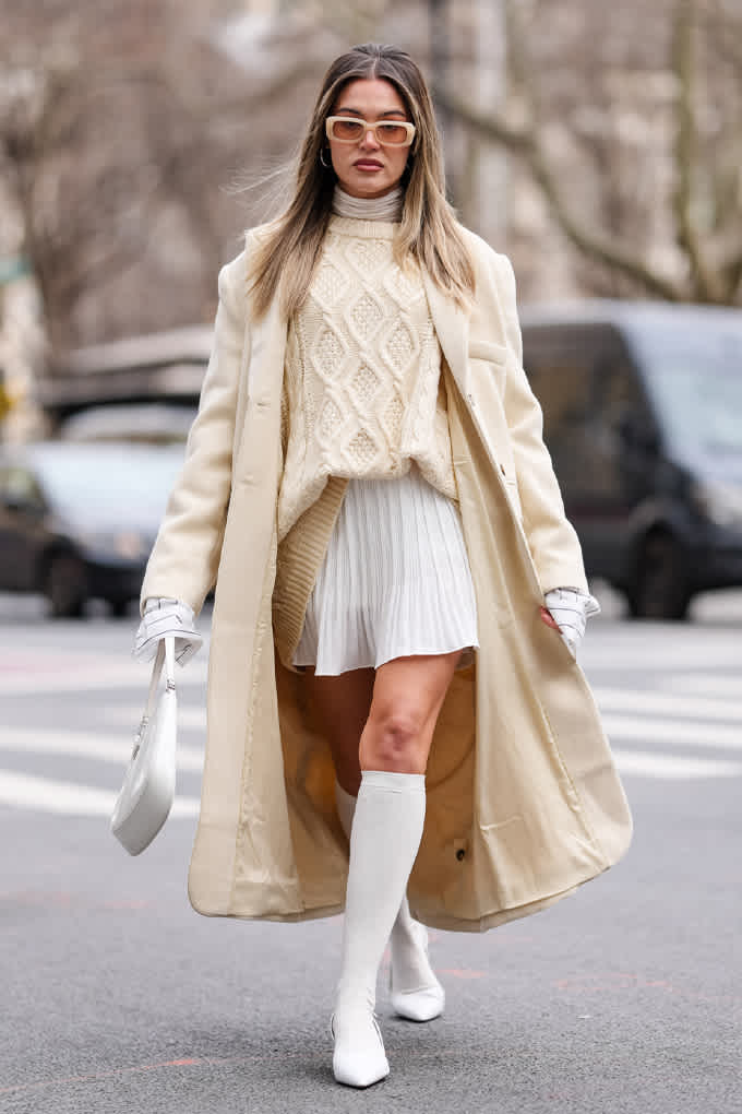 Frau in Oversize-Mantel mit Strick-Oberteil und Mini-Plissee-Rock, alles im Off-White-Look 