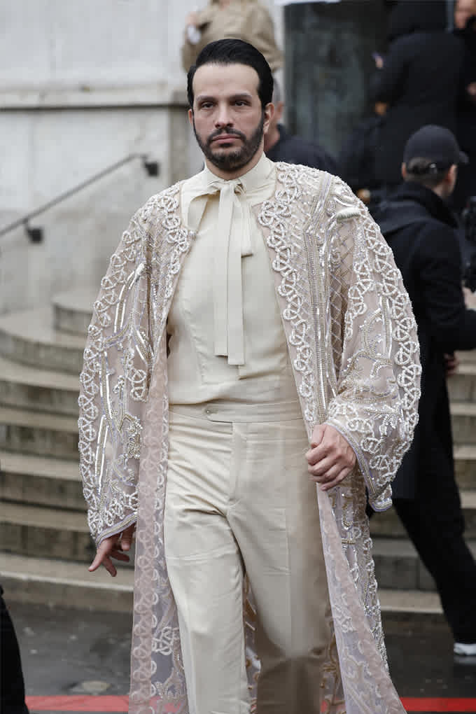 Mann in elegantem weißen Haute Couture-Outfit, kombiniert mit transparentem Überwurf mit Musterung