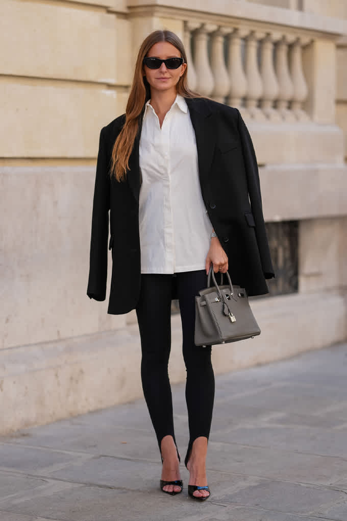 Una donna bionda indossa un blazer nero con logo abbinato a una camicetta bianca, leggings neri e una mini-borsa