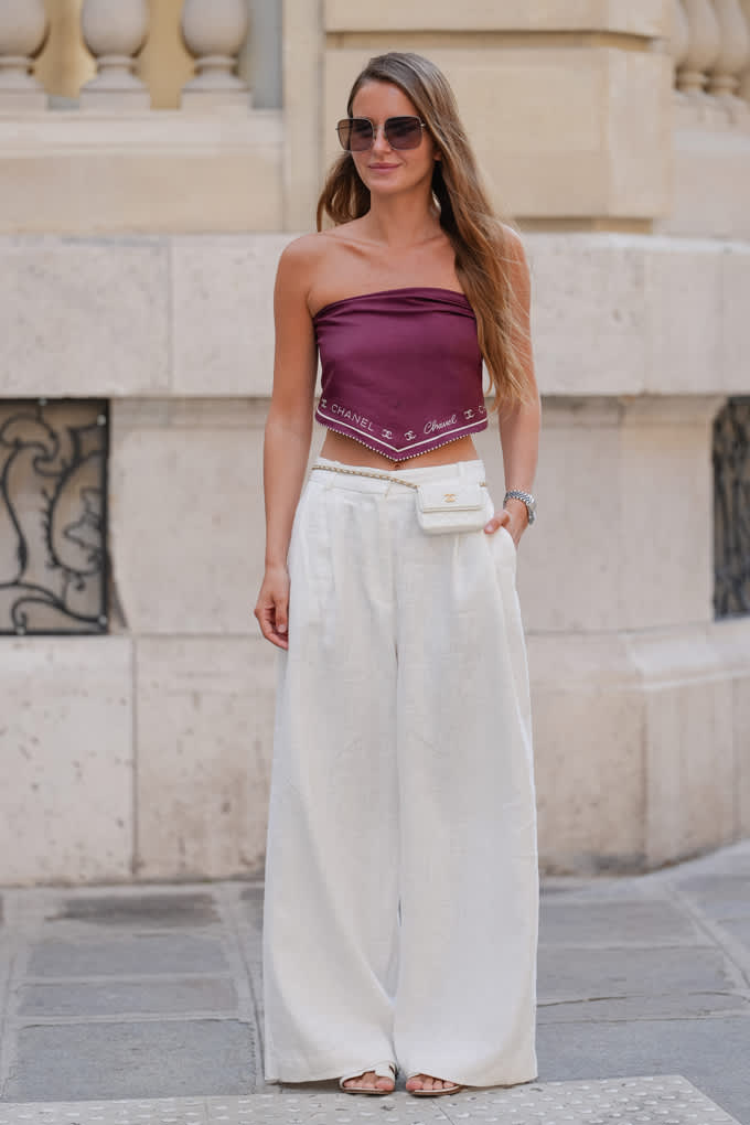 Donna con un top corto rosso e pantaloni di lino bianchi, in piedi davanti a un muro di pietra