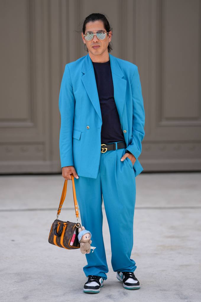 Mann kombiniert blauen Anzug mit schwarzem Shirt, Turnschuhen und Handtasche mit Plüsch-Anhänger