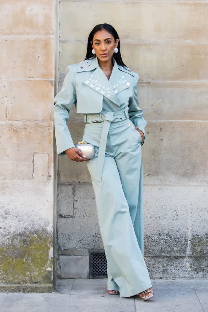 Frau in minzgrünem Blazer und passender Hose mit Clutch, lehnt entspannt an Steinwand