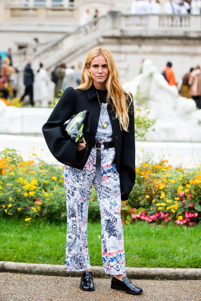 Una donna bionda indossa un blazer nero taglia XXL con pantaloni bianchi con motivi scarabocchiati e mocassini, in piedi davanti a un'area verde
