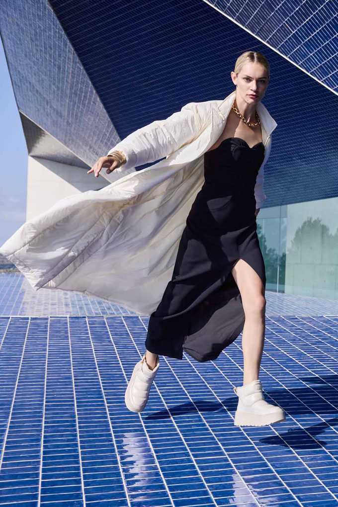 Eine Frau trägt ein schwarzes Kleid mit hohem Schlitz, kombiniert mit einem langen, weißen Mantel und weißen Boots