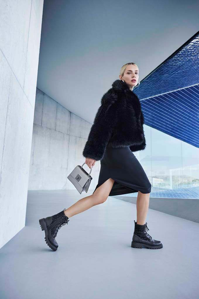 Eine Frau trägt ein schwarzes Kleid mit Schlitz, dazu eine schwarze Felljacke, klobige Boots und eine kleine graue Handtasche
