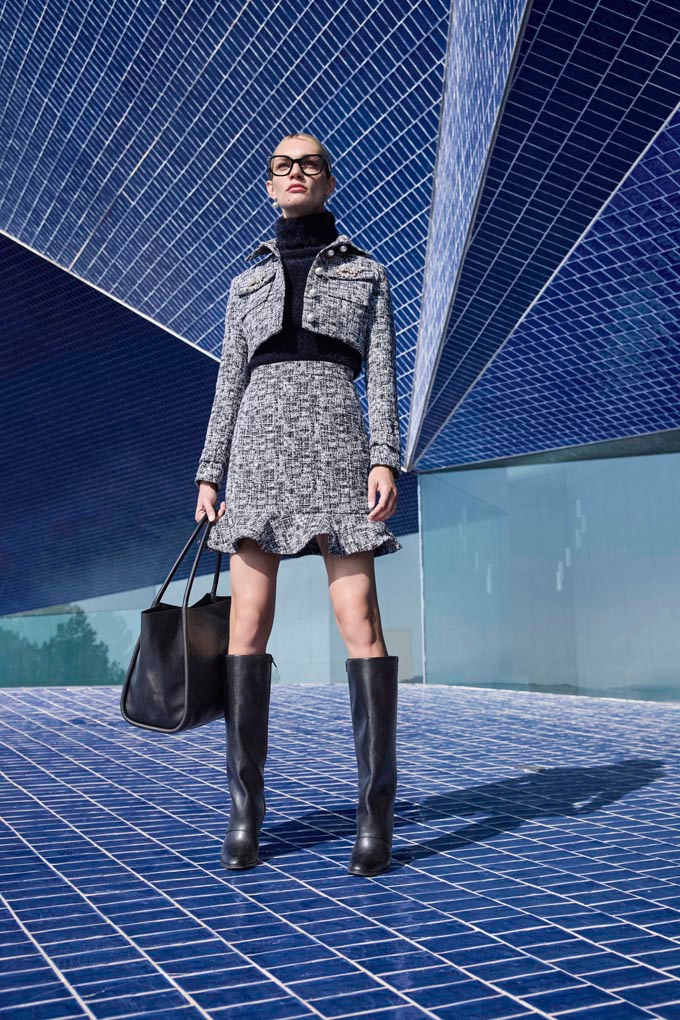 Eine Frau trägt einen grauen Rock, eine graue Jacke, schwarze kniehohe Stiefel und eine große schwarze Tasche