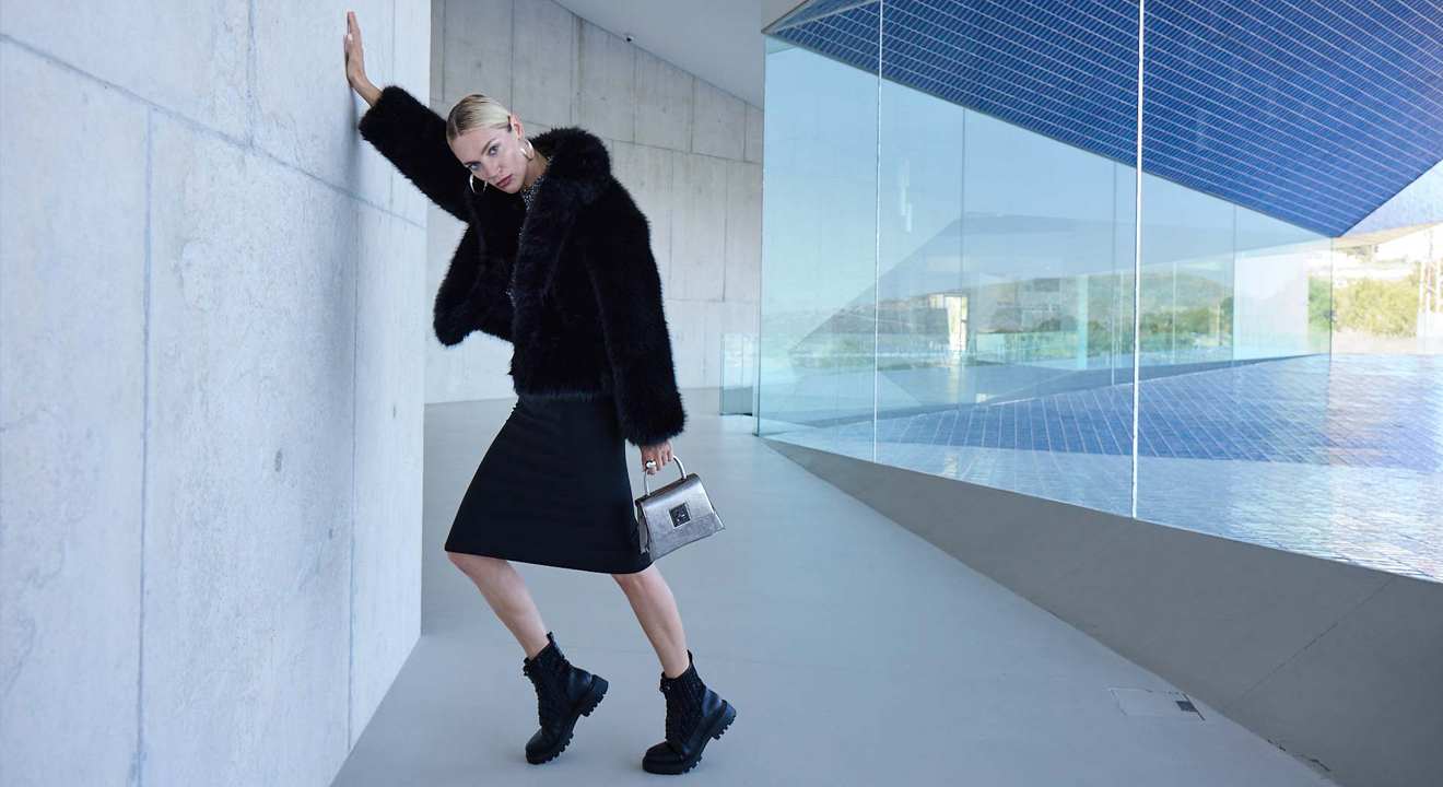 Eine Frau trägt einen schwarzen Kunstpelzmantel, einen schwarzen Rock und klobige Schnürboots; dazu kombiniert sie eine silberne Handtasche.