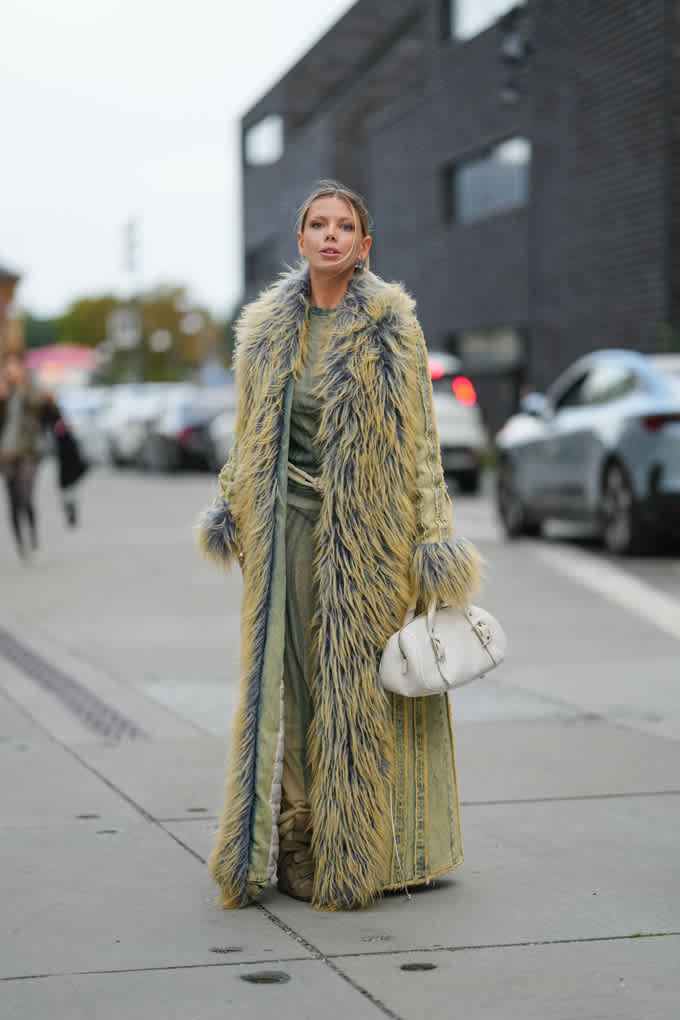 La donna indossa un lungo cappotto di pelliccia sintetica giallo-grigio, un completo abbinato, scarpe beige e una borsa bianca: un look appariscente e stravagante