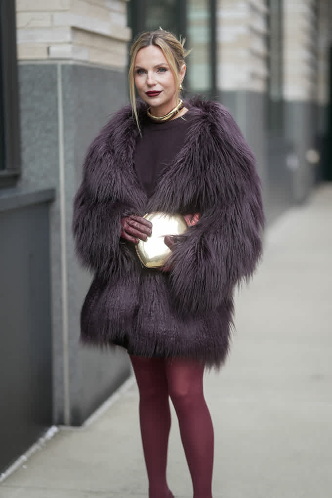 La donna indossa un cappotto di pelliccia sintetica viola con collant abbinati e una pochette dorata: un look elegante e coordinato