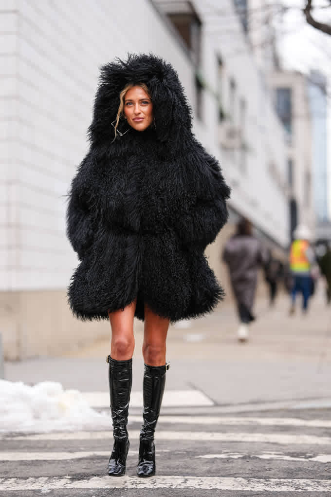 Una donna indossa un voluminoso cappotto di pelliccia nero con cappuccio e lo abbina a stivali di vernice lucidi al ginocchio