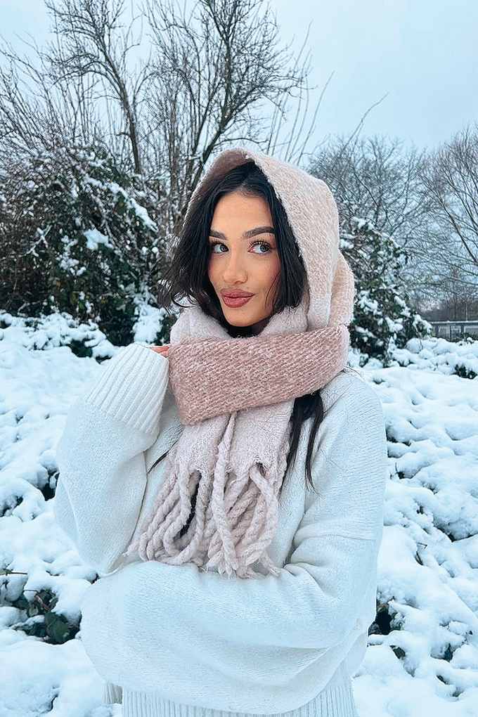 Rossella steht in weißem Pullover mit beigem Schal und Kapuze in Schnee-Kulisse
