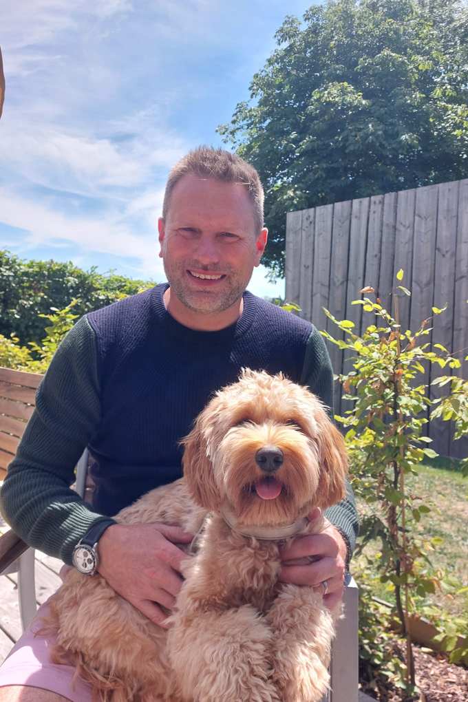 Un uomo con un maglione blu è seduto su una sedia da giardino, con un cane dal pelo soffice in grembo