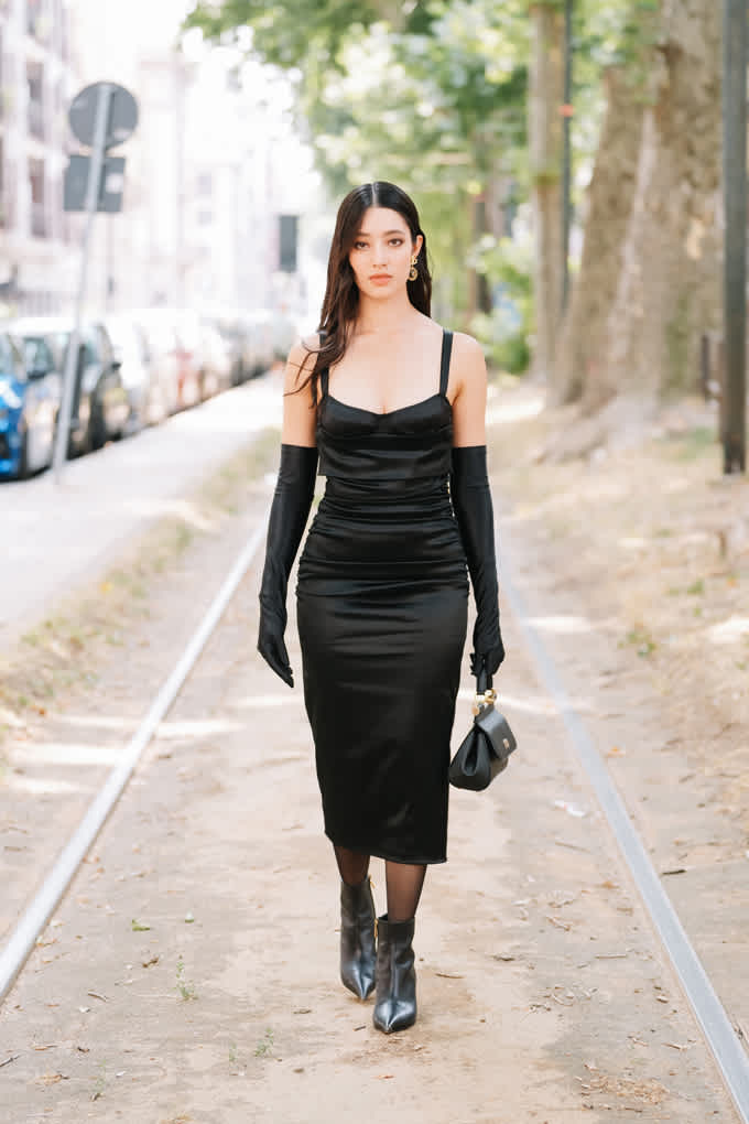 Schwarzhaarige Frau in schwarzem Satinkleid mit passenden Handschuhen, trägt kompakte Handtasche und Ankle Boots