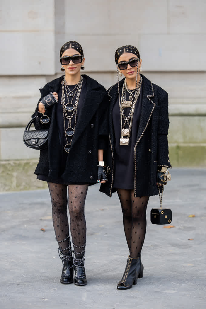 Zwei Frauen in modischen, schwarzen Outfits mit Oversize-Blazern, behangen mit Goldketten 