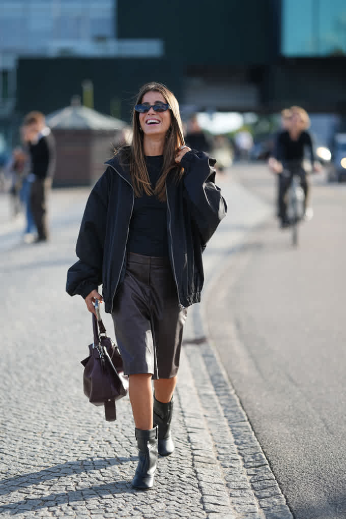 Lächelnde Frau mit schwarzer Jacke, trägt knielange Hose und Ankle Boots mit Blockabsatz