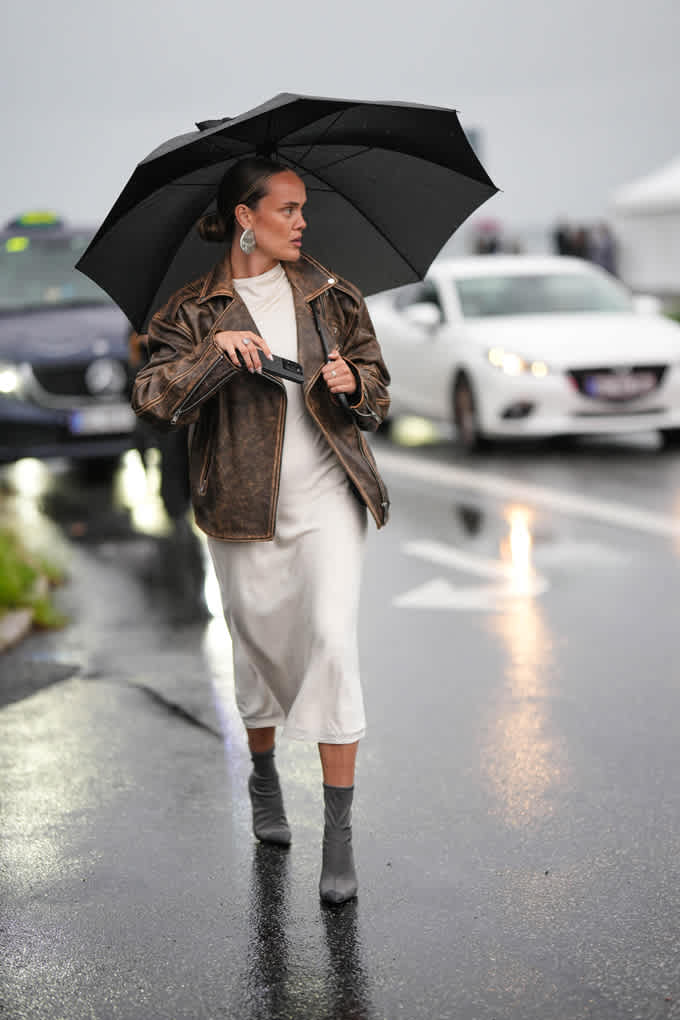 Frau trägt braune Lederjacke zu einem weißen Kleid, läuft mit Regenschirm an Straße entlang