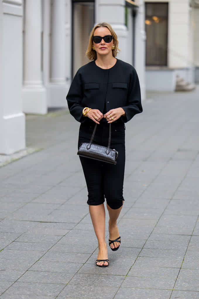 Frau in schwarzem Outfit auf Gehweg, trägt passende Baguette Bag in den Händen