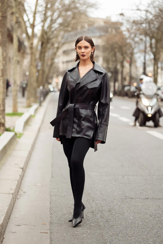 Frau mit strengem Blick und strammem Mittelscheitel, posiert auf Straße, trägt schwarzen Ledermantel und High Heels