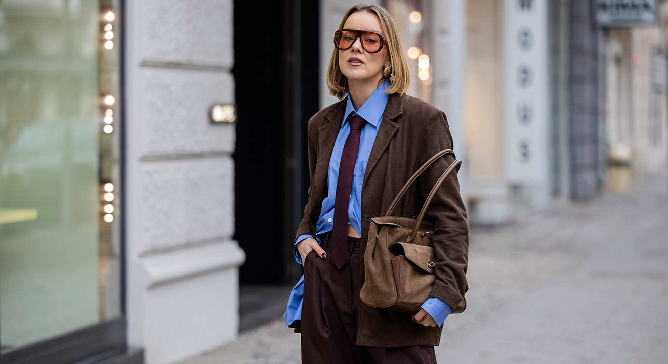 Blonde Frau trägt braunen Blazer mit passender Hose, blauem Hemd und Krawatte, im Preppy Look