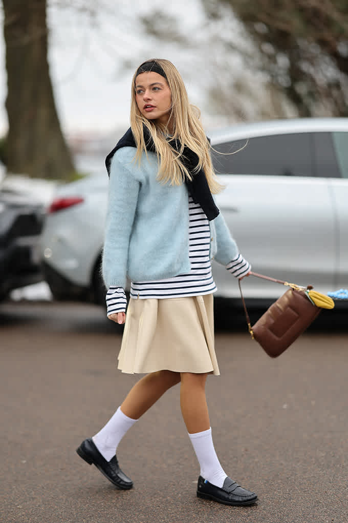Blonde Frau trägt hellblauen Cardigan über Streifenshirt, dazu Faltenrock und schwarze Loafer