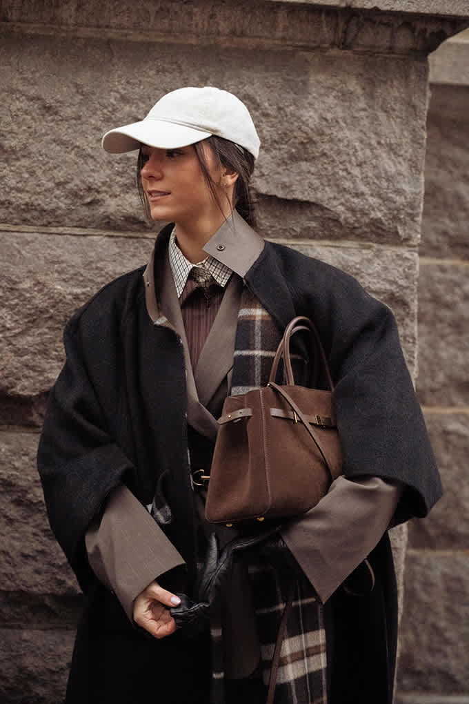 Frau in Tweed-Jacke, Karoschal und weißer Cap, steht vor Steinmauer