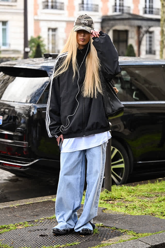 Frau mit lang-blonden Haaren, schwarzes XL-Jacket zu Baggy Pants, in Stadtkulisse