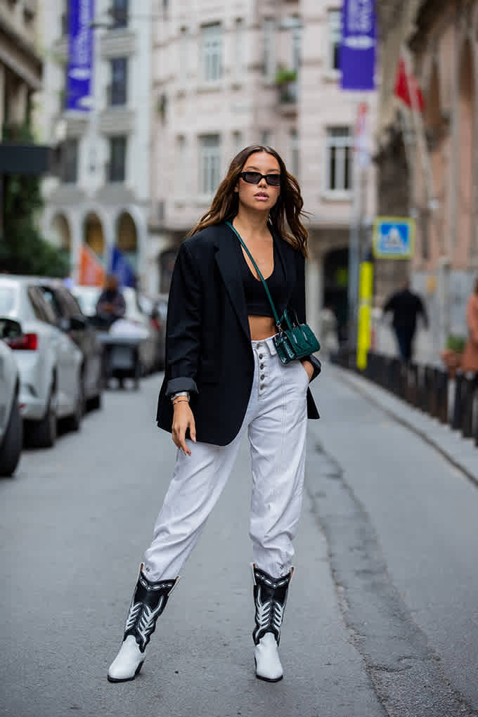 Frau steht auf Straße, trägt lockeren Blazer mit Tube Top um High-Waist-Pants mit weißen Cowboy Boots