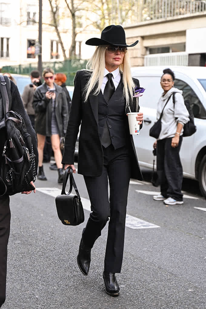 Frau mit blonden Haaren und schwarzem Anzug, mit weitem Hut und Boots