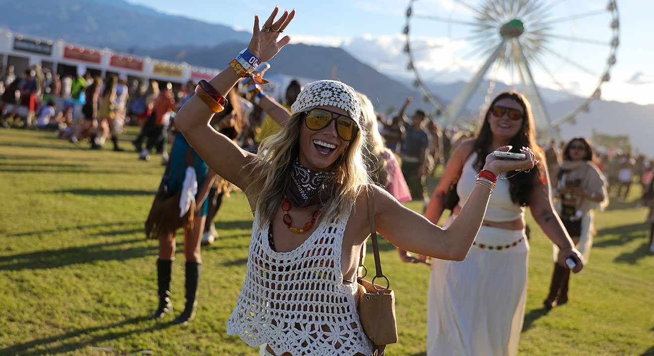 Eine fröhliche Frau im Coachella Outfit mit Häkeloberteil, Bandana und Sonnenbrille tanzt vor einem Riesenrad, während eine Begleiterin im weißen Maxirock hinter ihr posiert.