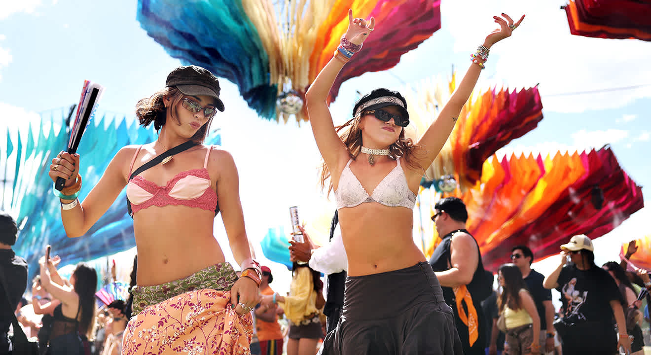 Zwei tanzende junge Frauen in typischen Coachella-Outfits (Bralettes, Sonnenbrillen und Schirmmützen) unter farbenfrohen Kunstinstallationen.