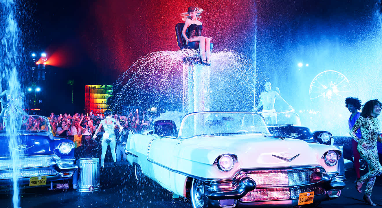 Sabrina Carpenter performt in einem glitzernden Top ( Coachella-Outfit) auf einer schwebenden Plattform über Oldtimern, umgeben von Wasserspielen und bunten Lichtern.