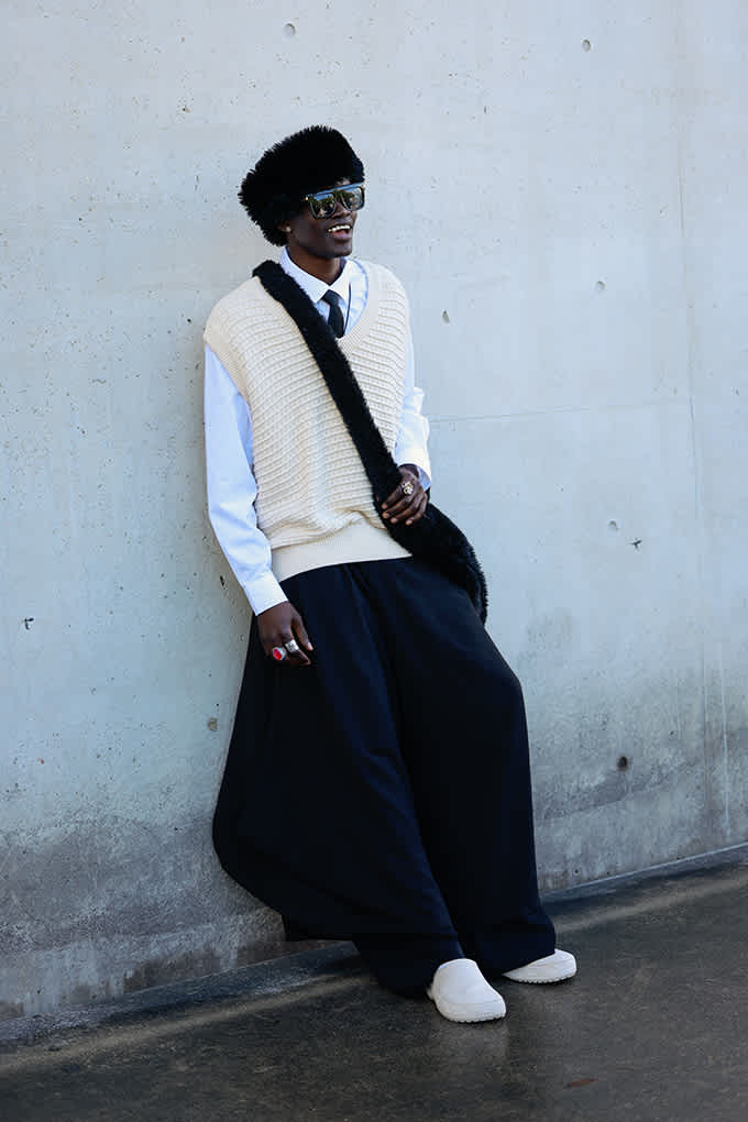 Schwarzer Mann in lässigem Outfit, trägt Pullunder, Krawatte und Wide-Leg-Hose, lehnt lächelnd an Wand