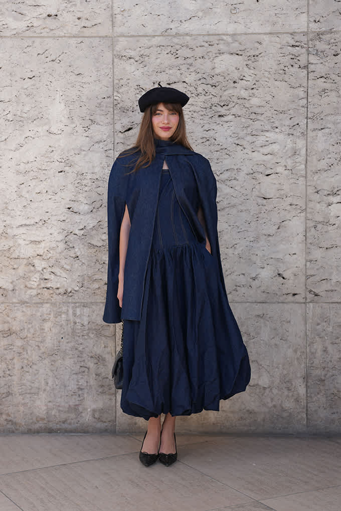 „Person in dunkelblauem Cape-Kleid und Baskenmütze vor heller Steinwand.