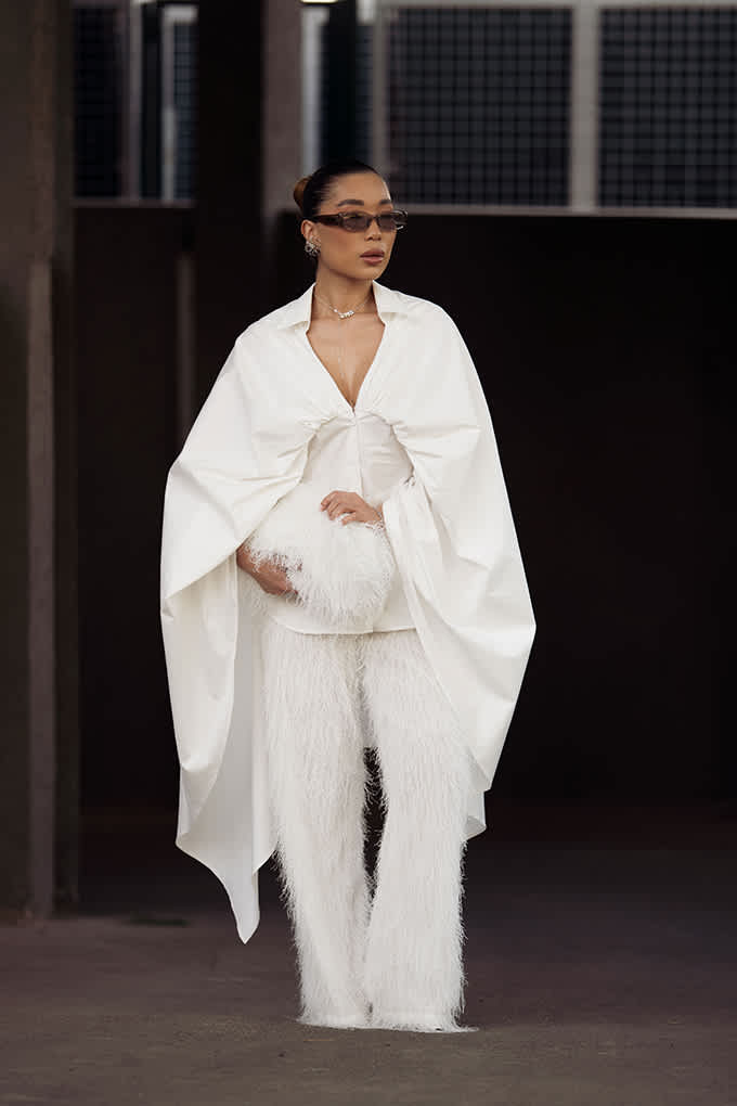 Person in weißem Cape-Top und strukturierter Hose mit Sonnenbrille und Clutch
