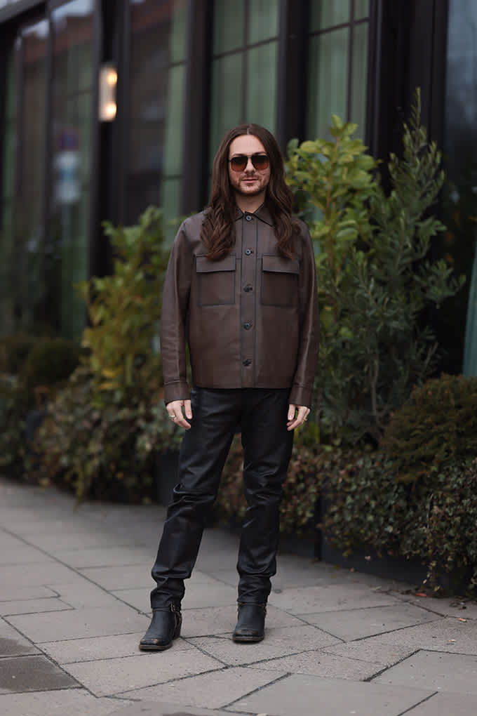Person in brauner Lederjacke und schwarzen Bikerboots vor einem modernen Gebäude