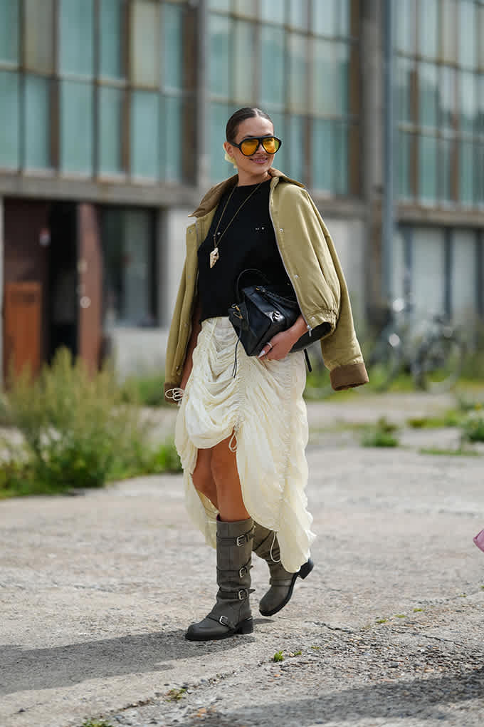 Frau läuft in kniehohen Bikerboots, hellem Rock und olivfarbener Jacke.