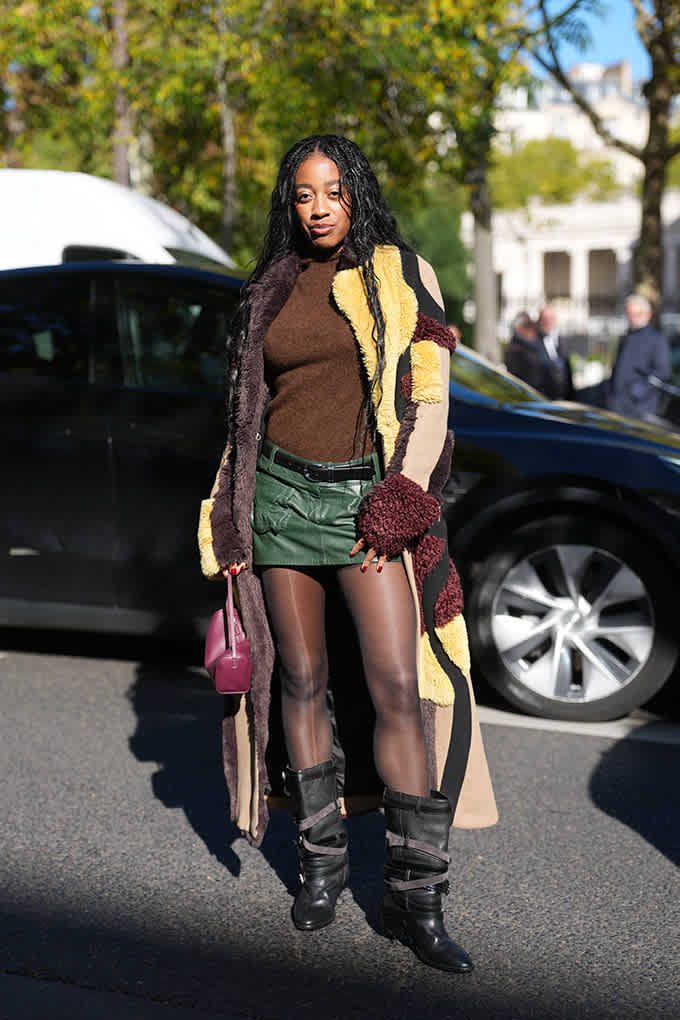 Frau steht vor einem Auto, trägt Patchwork-Mantel, grünen Leder-Minirock und Strappy Boots mit rockiger Ausstrahlung.