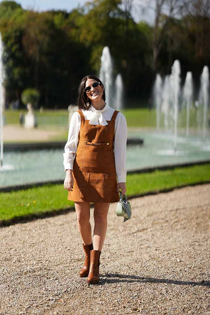 Junge Frau mit schwarzem Haar, trägt Suede-Pinafore-Mini über einer Bluse mit Schleife, vor Wasserspiel in Park