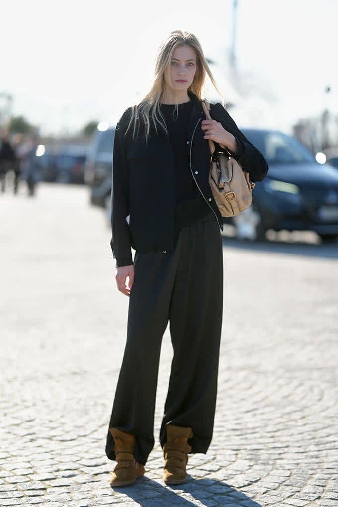 Eine Frau trägt ein schwarzes Outfit und eine beigefarbene Tasche. Besonders auffällig sind ihre braunen Sneaker-Wedges mit breiter, erhöhter Sohle und markanten Riemen.