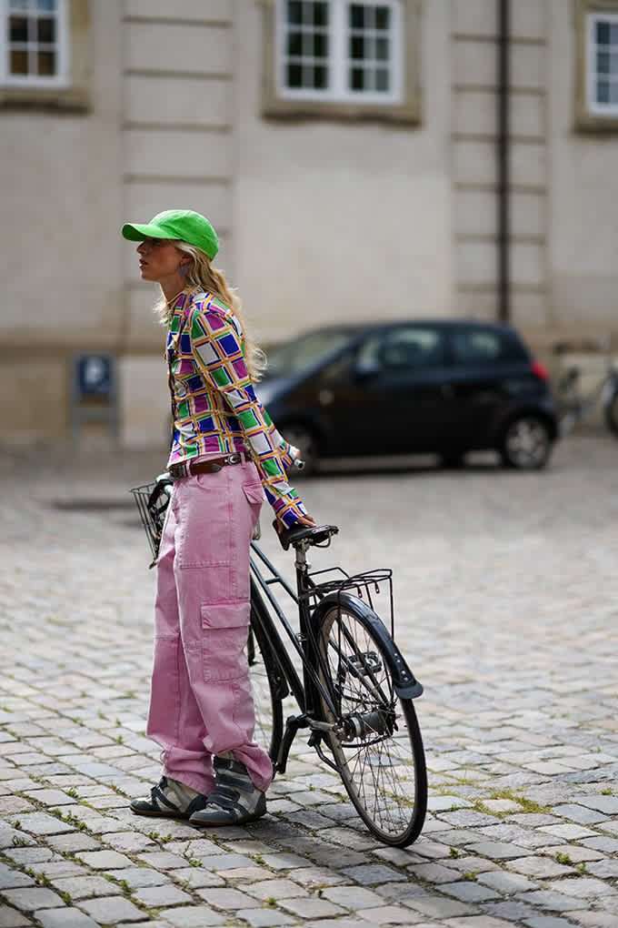 Eine Frau trägt ein bunt gemustertes Oberteil, eine grüne Cap und eine weit geschnittene rosa Cargohose. Dazu trägt sie graue Sneaker mit dunklen Details und einer robusten, leicht erhöhten Sohle.