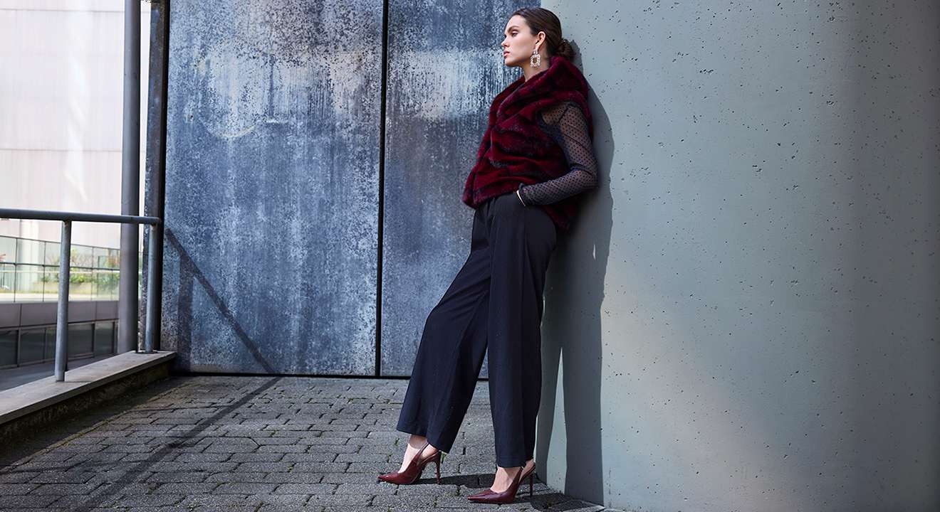 Eine Frau lehnt an einer Wand und trägt einen burgunderfarbenen Schal, eine schwarze Hose und rote Pumps.