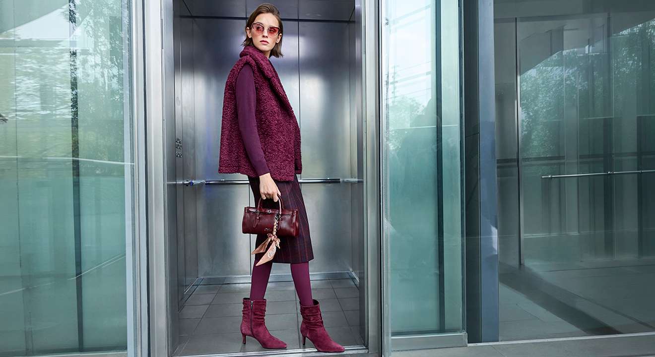 Eine stilvoll gekleidete Frau mit bordeauxrotem Outfit und Handtasche steht in einem gläsernen Aufzug.